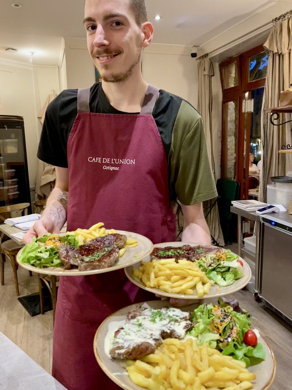 Un serveur en tablier bordeaux portant trois assiettes de viande, frites et salade